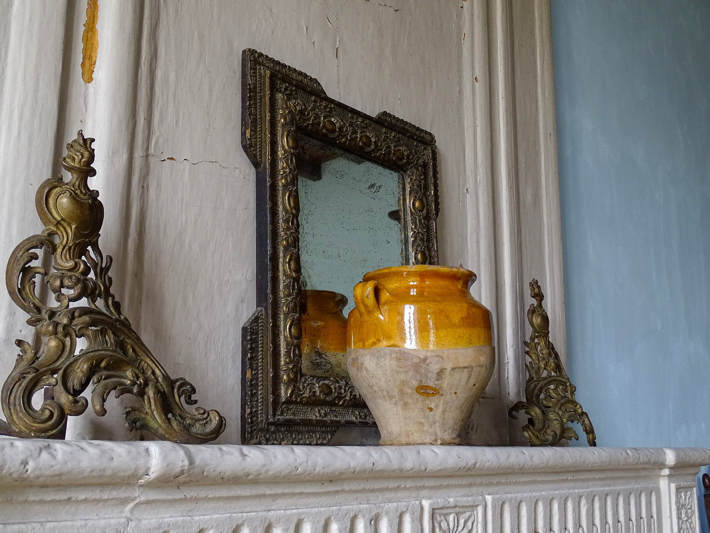 Antique French confit pot with yellow glaze on an old stone fireplace mantel in a French country house with an antique gold mirror and brass andirons.