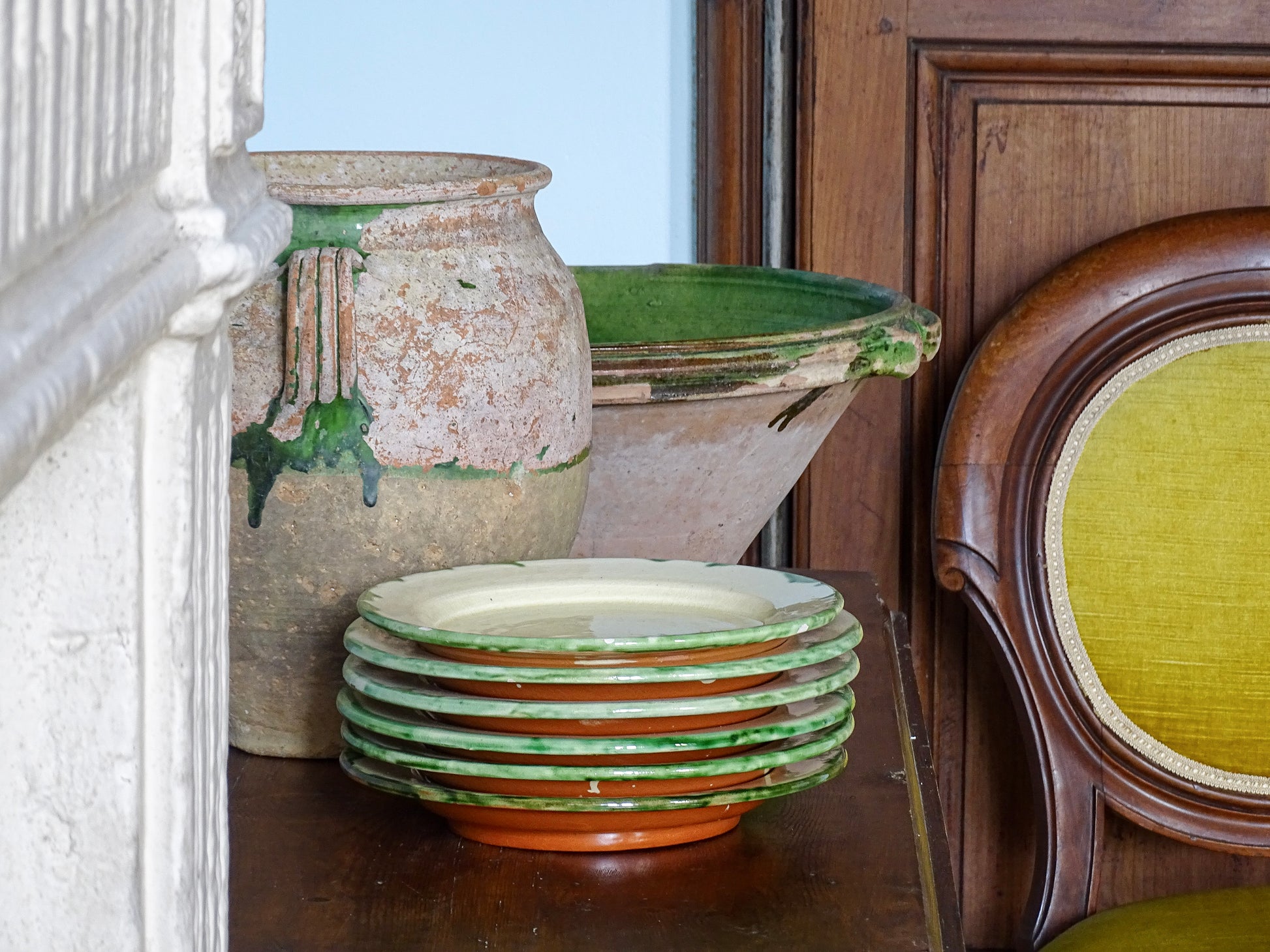 French vintage ceramic plates with green glaze trim alongside an antique confit pot and green glazed tian bowl next to an old stone fireplace, a large armoire and a chair with yellow velvet in a French country house.