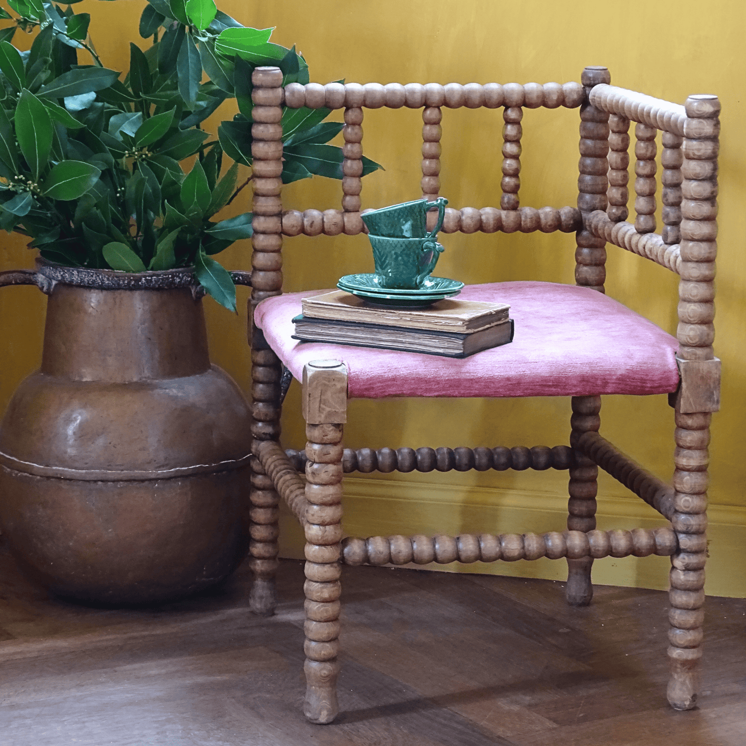 Antique wooden bobbin chair with pink cushion and green cup and books, next to a large copper pot with green plant against a mustard yellow wall.